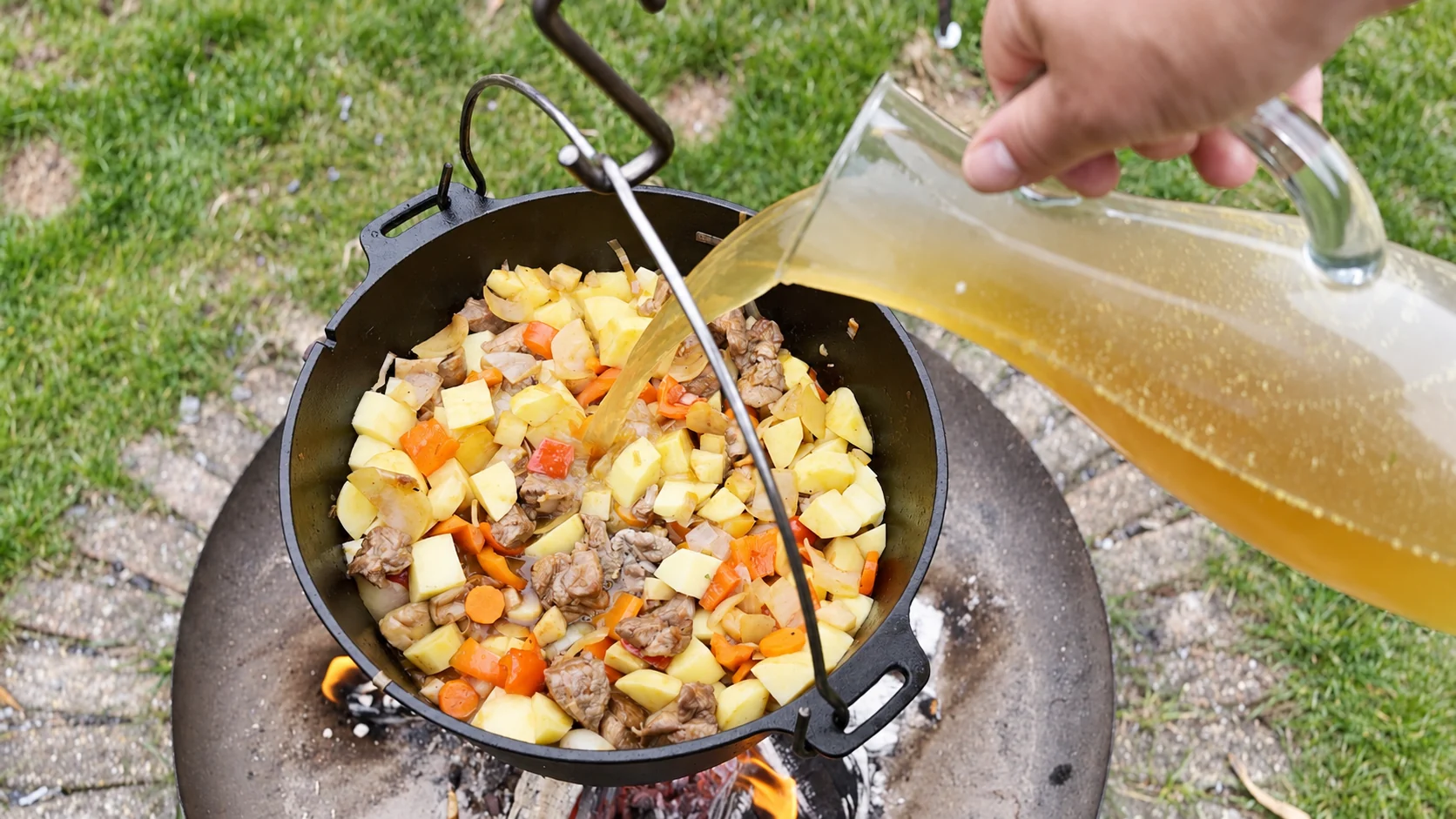 Schmorflüssigkeit wird aus einer Glaskanne zum Schweinegulasch, den Kartoffeln und dem Suppengemüse gegossen.
