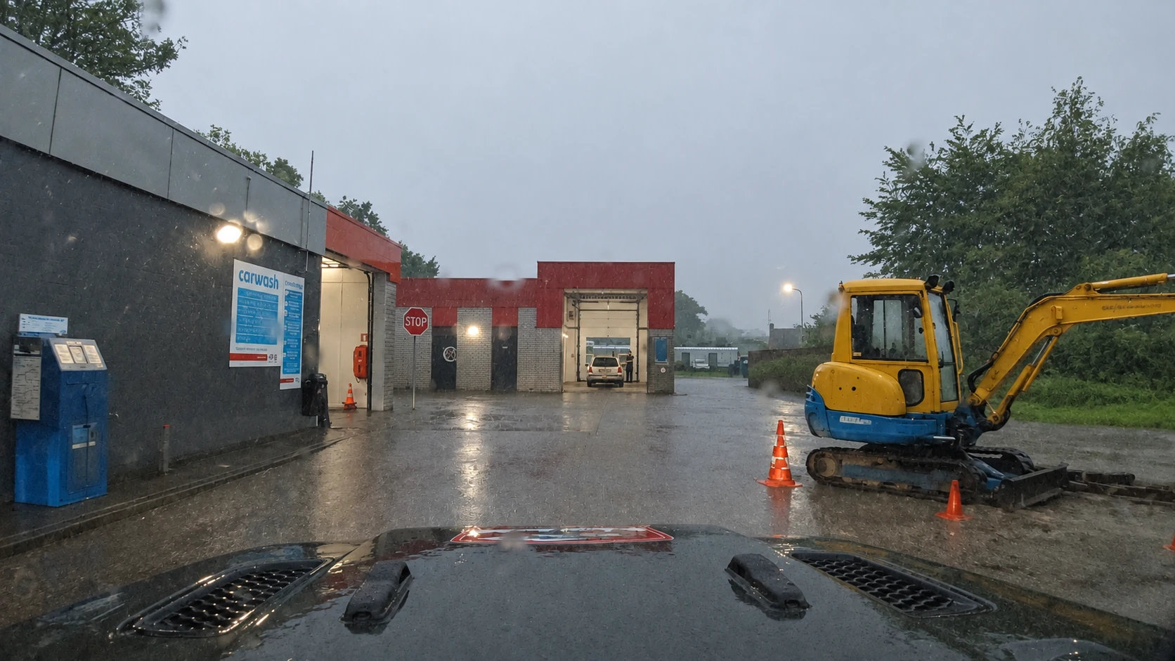 Regen und stehendes Wasser vor Waschhallen mit einem gelben Bagger rechts