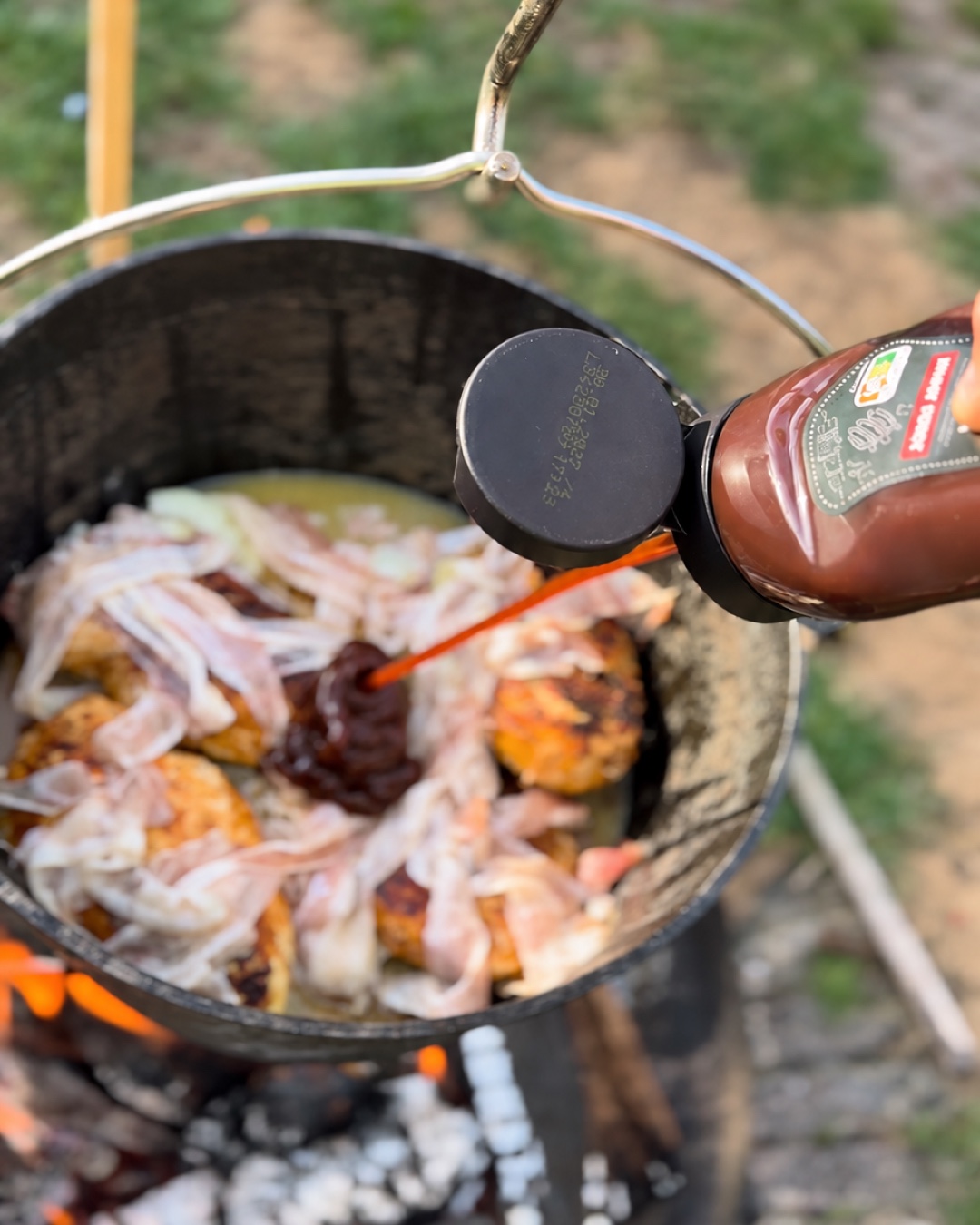 BBQ-Soße wird in den Dutch Oven zu Hähnchen, Bacon und Zwiebeln gegeben.