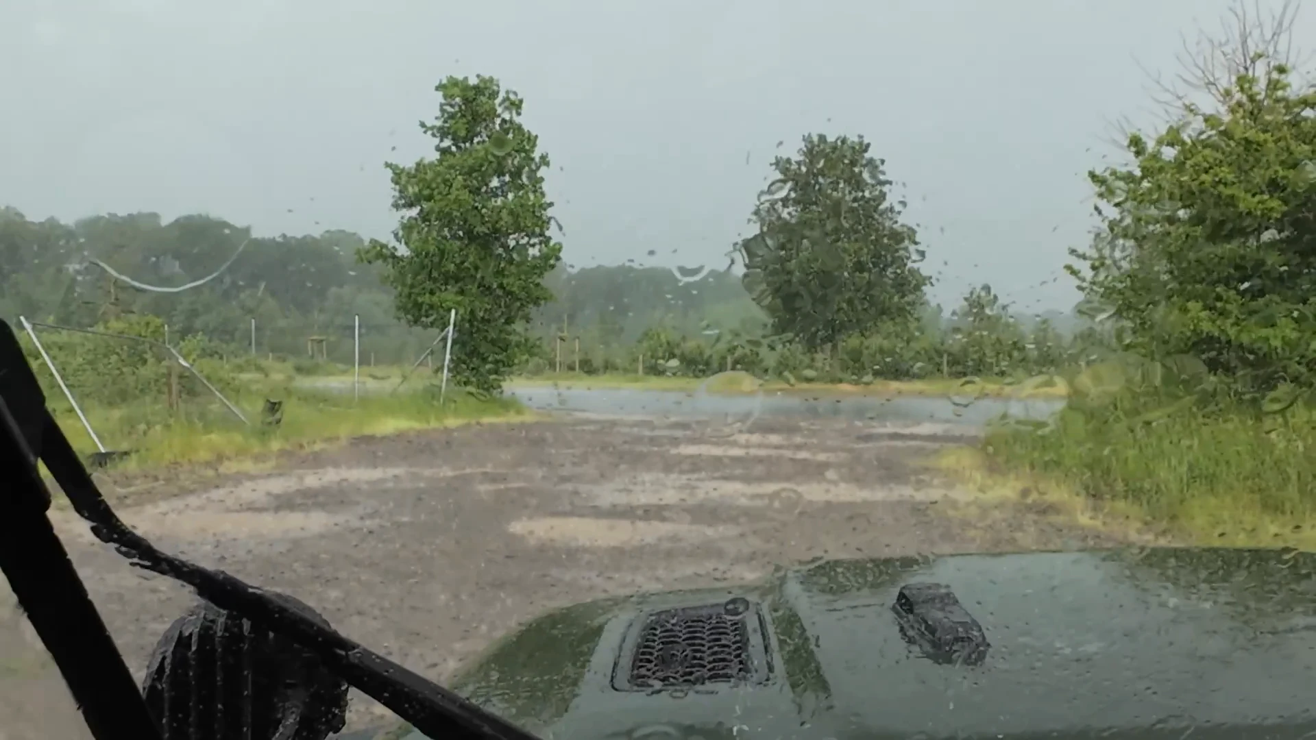 Blick aus dem Jeep durch die regennasse Frontscheibe auf einen Schotterplatz und grüne Bäume.