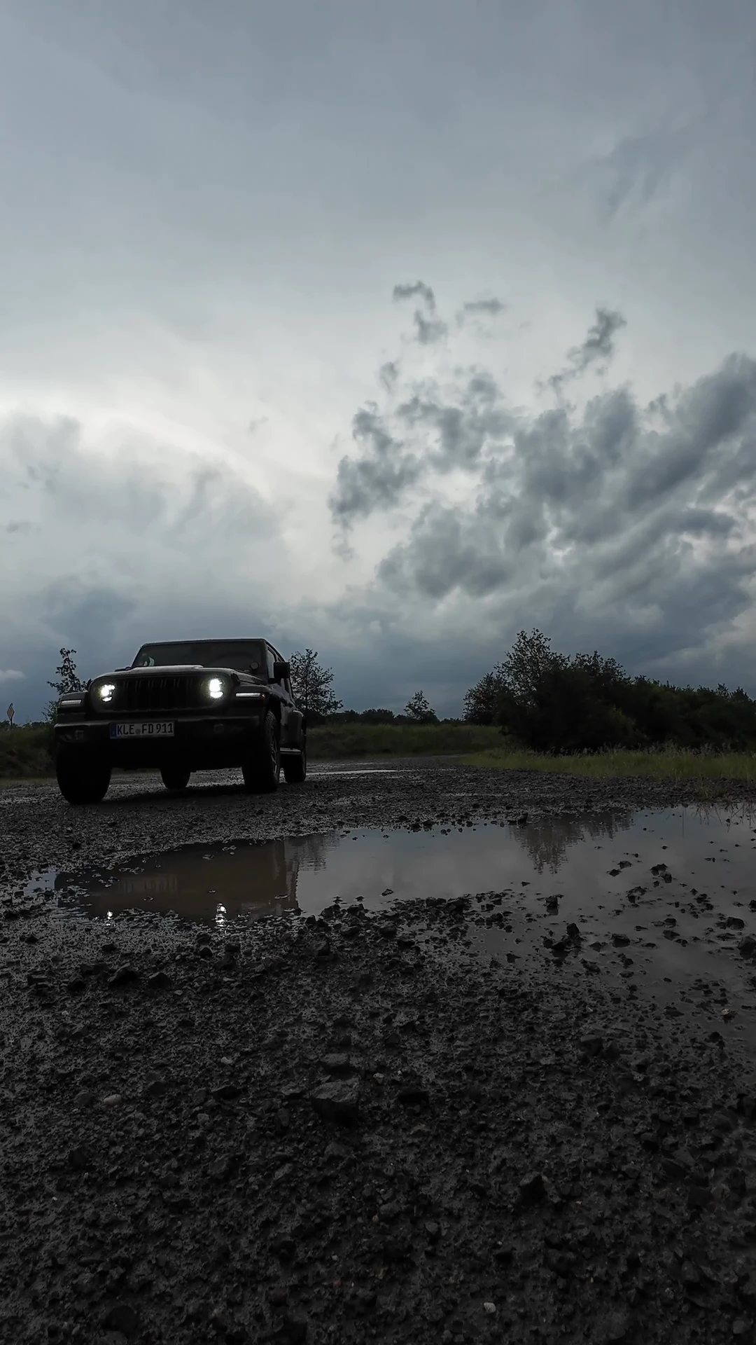Der Jeep mit eingeschalteten Scheinwerfern auf nassem Schotter neben einer Pfütze, in der sich die Lichter spiegeln.