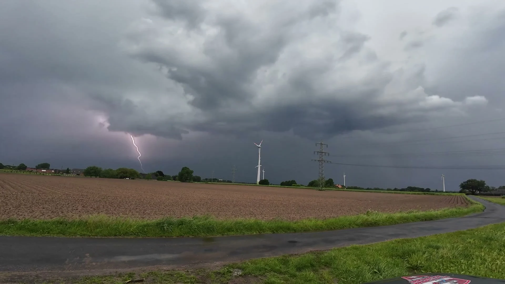 Ein heller Blitzkanal unter dunklen Wolken über einem Acker, rechts davon Windräder und ein Hochspannungsmast.