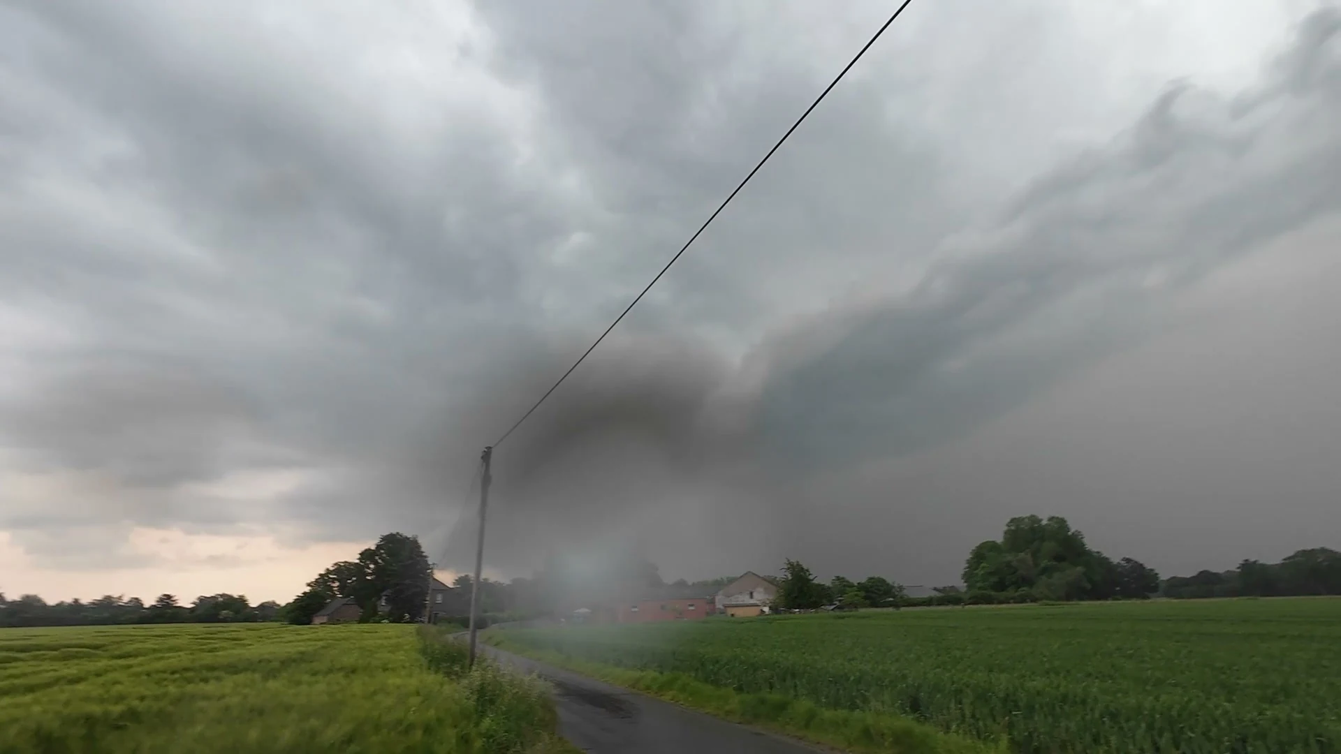 Dunkle Wolken über einem grünen Feld und einer schmalen Straße; unter den Wolken liegt ein grauer Niederschlagsvorhang.