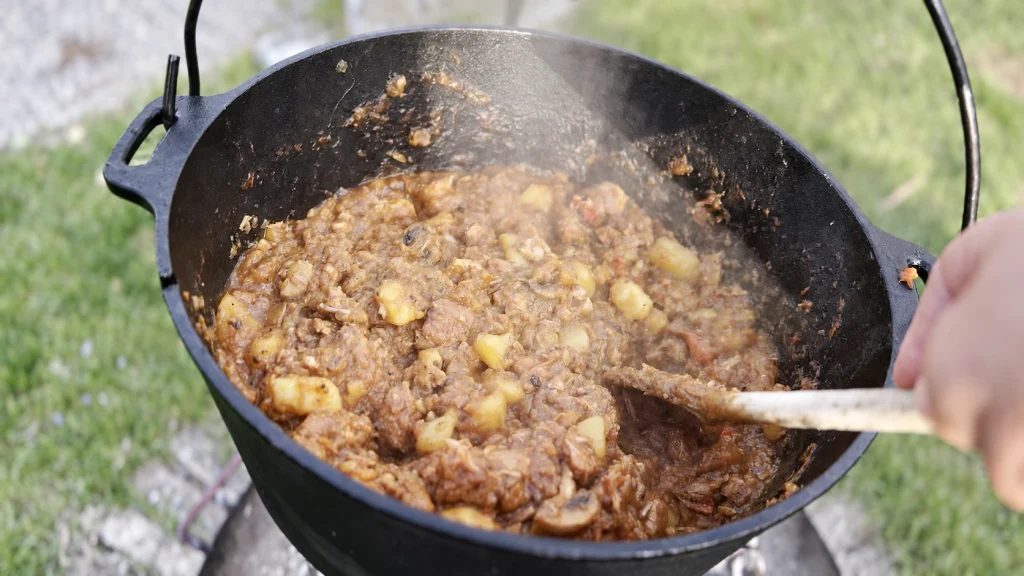 Schweinegulasch, Kartoffeln und Gemüse im Dutch Oven, mit einem Holzlöffel umgerührt.